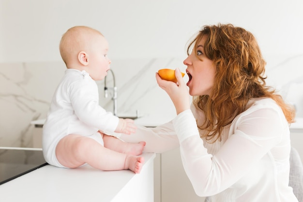 Retrato de uma jovem mãe sentada e comendo laranja enquanto brincava com seu bebezinho na cozinha