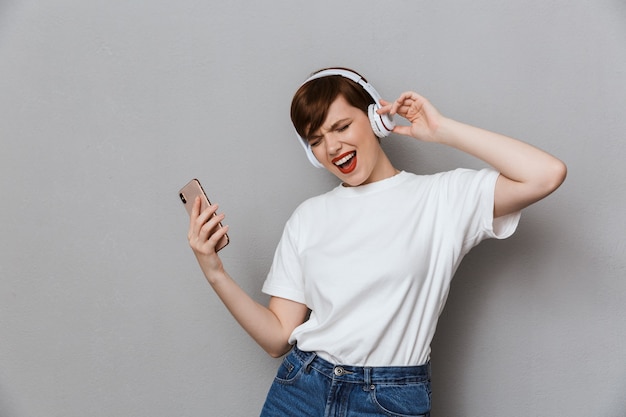 Retrato de uma jovem maravilhada cantando enquanto ouve música com fones de ouvido e smartphone isolados sobre uma parede cinza