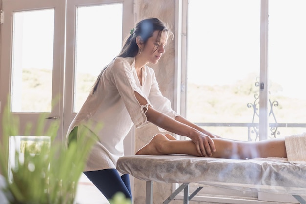 Retrato de uma jovem massagista realizando uma massagem nas pernas em uma mulher deitada em uma mesa de massagem Vista lateral