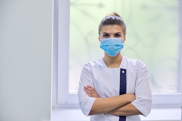 Retrato de uma jovem médica confiante em uma máscara médica com os braços cruzados sobre os interiores da clínica, copie o espaço