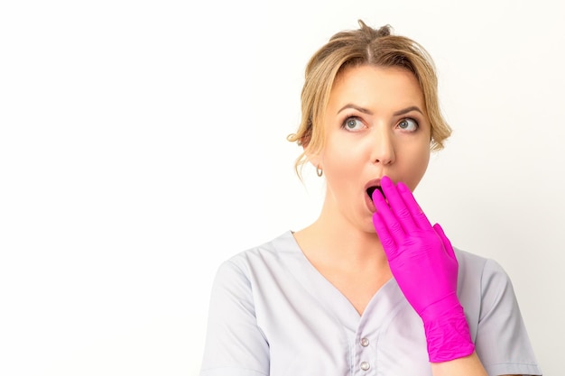 Retrato de uma jovem médica ou enfermeira caucasiana fica chocada cobrindo a boca com as mãos enluvadas rosa contra um fundo branco