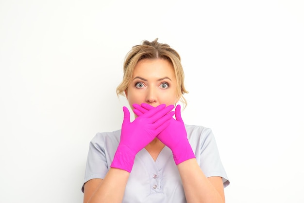 Retrato de uma jovem médica ou enfermeira caucasiana fica chocada cobrindo a boca com as mãos enluvadas rosa contra um fundo branco