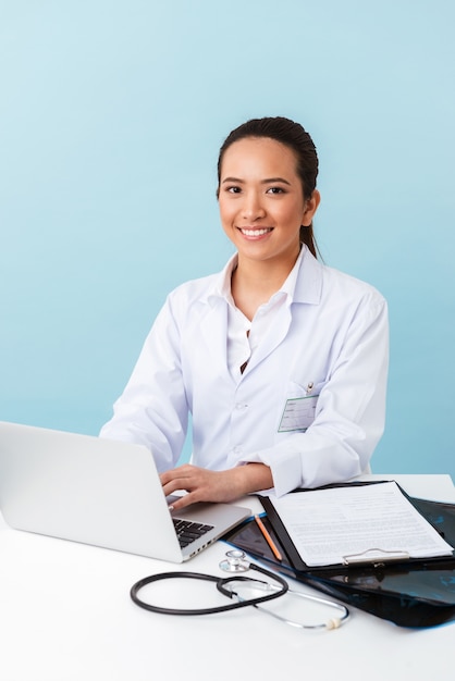 Foto retrato de uma jovem médica posando isolado sobre uma parede azul, usando o computador portátil.