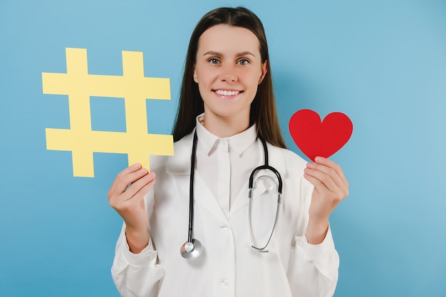 Retrato de uma jovem médica simpática com sorriso dentuço e bom humor segurando uma grande placa de hashtag amarela e um coraçãozinho vermelho, usa um casaco, posando isolado sobre um fundo de cor azul no estúdio