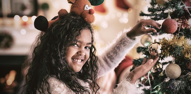 Retrato de uma jovem mestiça em sua sala de estar no Natal, usando um chapéu festivo segurando uma bugiganga em uma árvore de Natal sorrindo