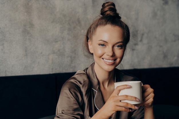 Retrato de uma jovem morena atraente com um lindo sorriso largo bebendo café na cama