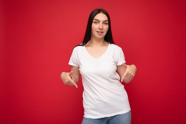 Retrato de uma jovem morena atraente positiva com emoções sinceras, vestindo camiseta branca casual para maquete isolada sobre fundo vermelho com espaço de cópia