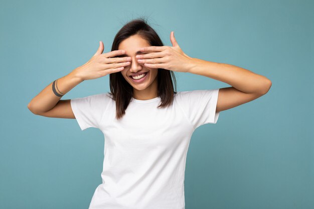 Retrato de uma jovem morena bonita feliz positiva com emoções sinceras, vestindo uma camiseta branca casual para maquete isolada em um fundo azul com um espaço vazio e cobrindo os olhos com as mãos