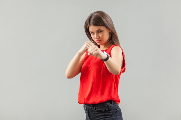 Retrato de uma jovem morena bonita séria em pé de camisa vermelha com punhos de boxe e olhando para a câmera, pronta para atacar ou defender. interior, tiro de estúdio, isolado em fundo cinza.