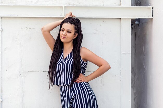 Retrato de uma jovem morena feliz com penteado preto afro americano dreadlocks em vestido listrado em pé, posando e olhando para longe e sorrindo. modelo de moda tiro.