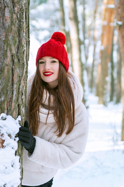 Retrato de uma jovem morena feliz sorridente caucasiana olhando atrás de uma árvore em uma floresta de inverno nevado de coníferas. Ao ar livre. Quadro vertical.