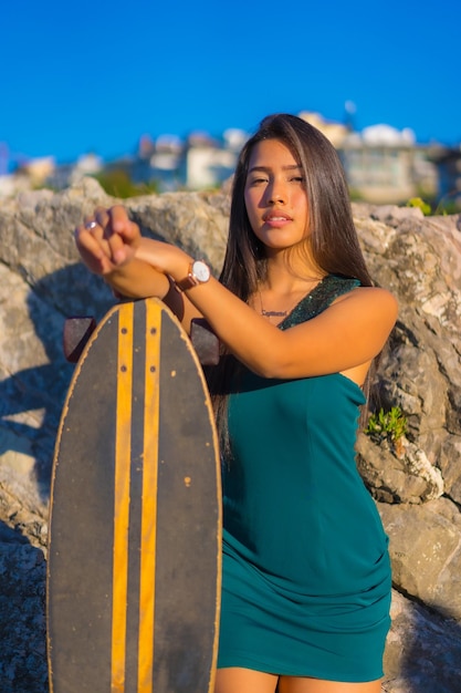 Retrato de uma jovem morena latina com um skate usando um vestido verde olhando para a câmera