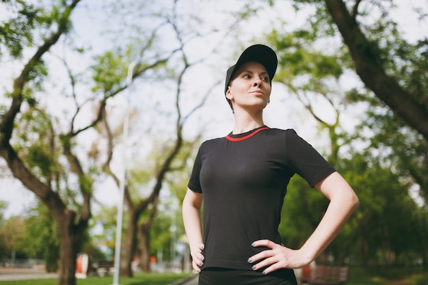 Retrato de uma jovem morena linda atlética de uniforme preto e boné em pé, descansando e olhando para o lado depois de correr, treinar no parque da cidade ao ar livre