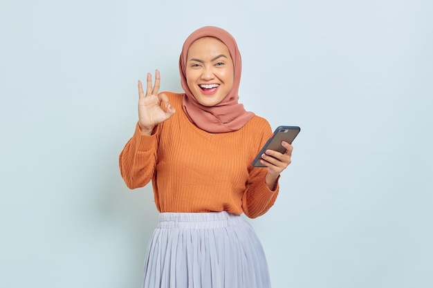 Retrato de uma jovem muçulmana asiática alegre de suéter marrom e hijab usando telefone celular fazendo gesto okey isolado no conceito de estilo de vida muçulmano de fundo branco