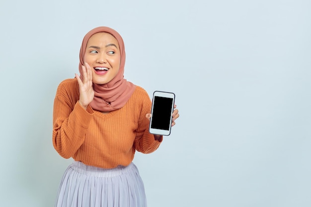 Retrato de uma jovem muçulmana asiática alegre de suéter marrom e lenço na cabeça mostrando a tela do celular em branco fazendo gesto de sussurro isolado no conceito de estilo de vida muçulmano de fundo branco