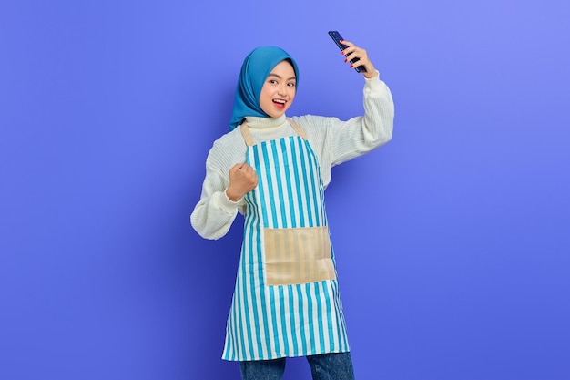 Retrato de uma jovem muçulmana asiática animada em 20 anos usando hijab e avental segurando o celular com gesto de vencedor isolado no fundo roxo. Conceito de estilo de vida muçulmano de dona de casa de pessoas