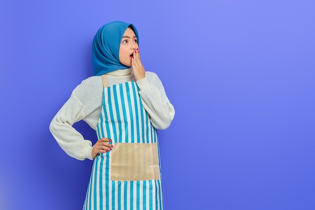 Retrato de uma jovem muçulmana asiática chocada em hijab e avental listrado cobrindo a boca com a mão olhando de lado isolado no fundo roxo Pessoas dona de casa conceito de estilo de vida muçulmano
