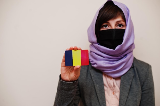 Retrato de uma jovem muçulmana vestindo roupa formal protege a máscara facial e o lenço de cabeça hijab segura o cartão da bandeira do chade contra o fundo isolado conceito de país coronavirus