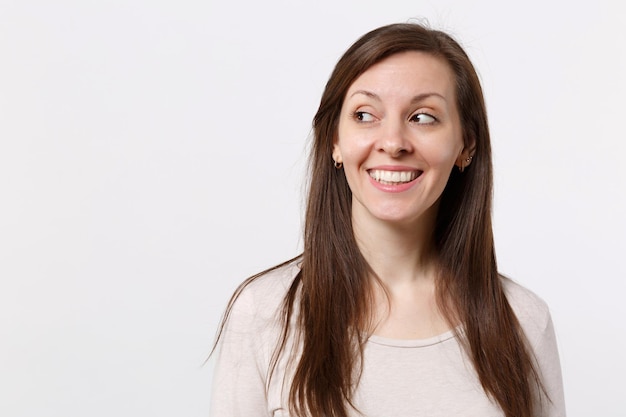 Retrato de uma jovem muito alegre sorridente em roupas leves em pé e olhando de lado isolado no fundo da parede branca no estúdio. Conceito de estilo de vida de emoções sinceras de pessoas. Mock up espaço de cópia.