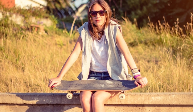 Retrato de uma jovem muito bonita com shorts curtos e skate, sentado na parede num dia de verão. Edição de tons quentes.