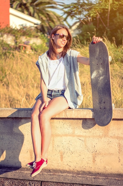 Retrato de uma jovem muito bonita com shorts curtos e skate, sentado na parede num dia de verão. Edição de tons quentes.