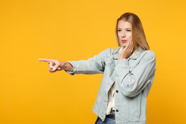 Retrato de uma jovem muito jovem em roupas casuais jeans, apontando o dedo indicador de lado, colocando a mão na bochecha isolada em fundo amarelo laranja no estúdio. Conceito de estilo de vida de pessoas. Mock up espaço de cópia.