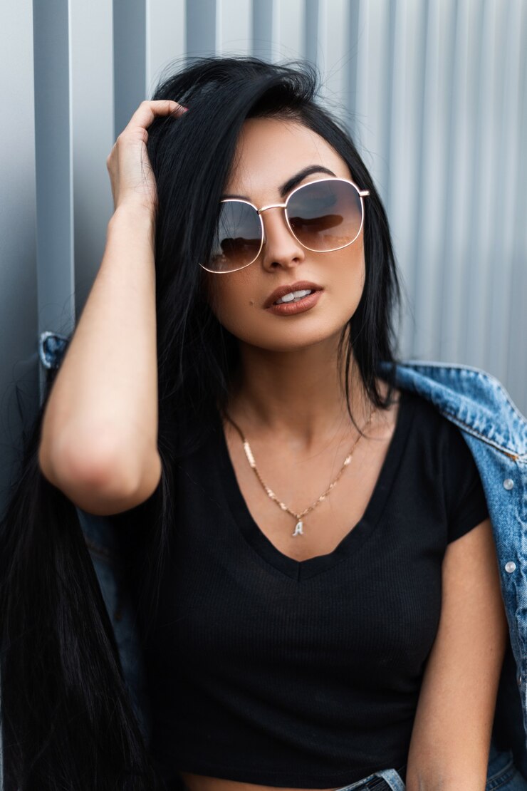 Foto retrato de uma jovem muito sexy com cabelo preto comprido luxuoso em uma elegante camiseta em óculos de sol da moda perto de uma parede de metal prata na cidade. garota atraente hipster é relaxante na rua.
