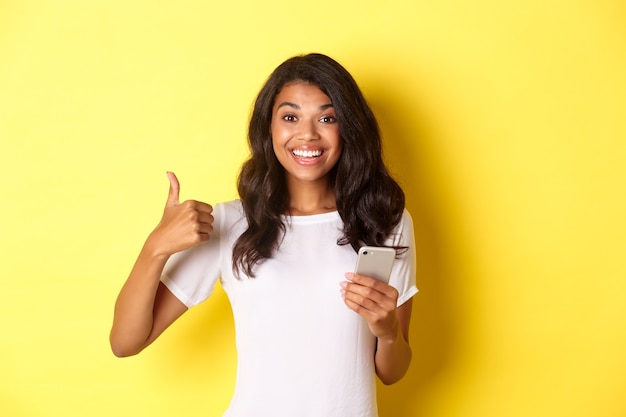 Retrato de uma jovem mulher afro-americana bonita, mostrando o polegar para cima ao usar o app do telefone móvel.