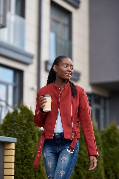 Retrato de uma jovem mulher afro-americana caminhando na rua e segurando uma xícara de café ao ar livre