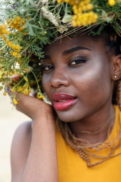 Retrato de uma jovem mulher afro-americana, modelo da moda, com grandes flores no cabelo. Retrato de uma menina em um plano torto em um campo com flores. coroa na cabeça dela