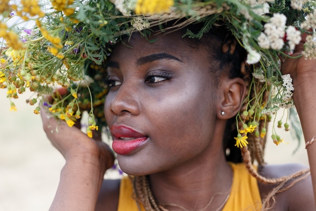 Retrato de uma jovem mulher afro-americana, modelo da moda, com grandes flores no cabelo. Retrato de uma menina em um plano torto em um campo com flores. coroa na cabeça dela