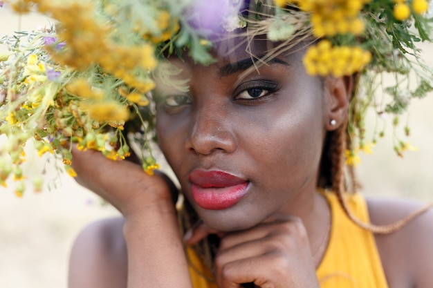 Retrato de uma jovem mulher afro-americana, modelo da moda, com grandes flores no cabelo. retrato de uma menina em um plano torto em um campo com flores. coroa na cabeça dela