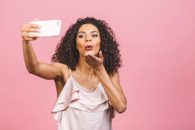 Retrato de uma jovem mulher afro-americana no vestido enviando beijo no ar em pé e tomando uma selfie isolada sobre fundo rosa.