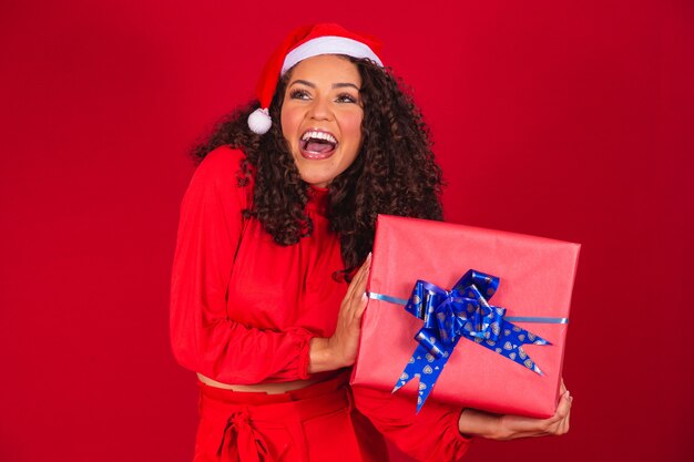 Retrato de uma jovem mulher afro com chapéu de Papai Noel segurando um presente sobre fundo vermelho. conceito de noite de natal