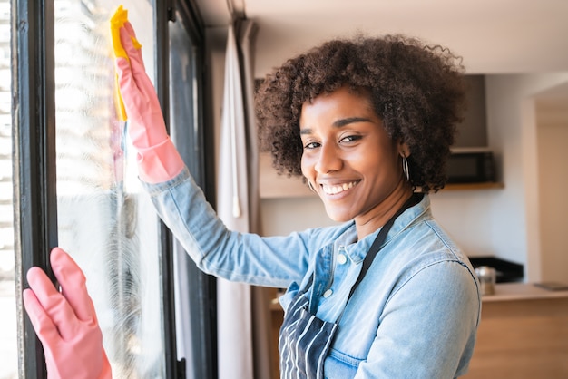 Retrato de uma jovem mulher afro em luvas, limpando a janela com um pano em casa. Conceito de trabalho doméstico, limpeza e limpeza.