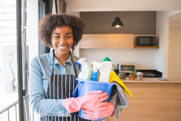 Retrato de uma jovem mulher afro, segurando um balde com produtos de limpeza em casa. conceito de arrumação e limpeza.