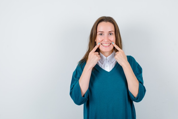 Retrato de uma jovem mulher apontando para as bochechas com um suéter por cima da camisa e olhando alegre para a frente