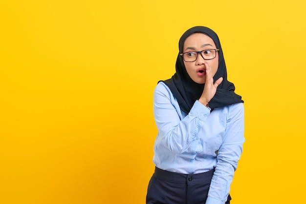 Retrato de uma jovem mulher asiática com a mão na boca contando um segredo, sussurrando uma fofoca isolada sobre fundo amarelo