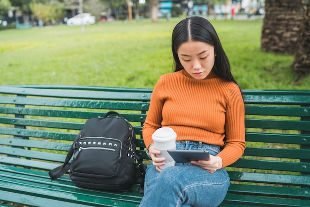 Retrato de uma jovem mulher asiática usando seu tablet digital enquanto segura uma xícara de café ao ar livre do parque.