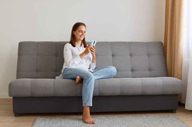 Retrato de uma jovem mulher atraente adulto segurando o telefone celular, verificando as redes sociais, olhando para a tela do telefone inteligente com um sorriso agradável, navegando na internet ou digitando mensagem.