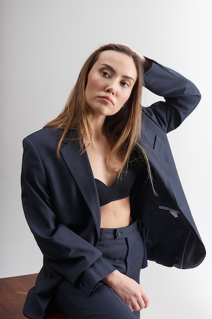 Retrato de uma jovem mulher atraente asiática com cabelo comprido no sutiã, azul terno sentado no banquinho. magro elegante muito feminino posando na parede branca. testes de modelo de bela senhora