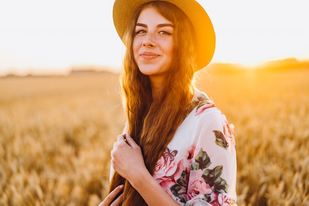 Retrato de uma jovem mulher bonita com cabelo encaracolado e sardas. Mulher de vestido e chapéu posando no campo de trigo ao pôr do sol e olhando
