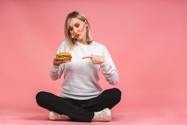 Retrato de uma jovem mulher bonita com fome comendo sanduíche de croissant. Retrato isolado de mulher com fast food sobre fundo rosa. Conceito de dieta. Sentado no chão em posição de lótus.