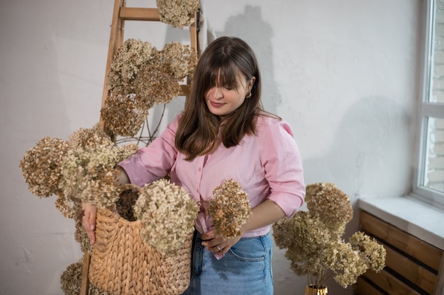 Retrato de uma jovem mulher bonita de cabelos escuros, florista no trabalho, fazendo buquês de hortênsias secas estabilizadas em cesta de vime.