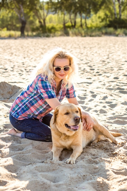 Retrato de uma jovem mulher bonita em óculos de sol, sentado na praia de areia com uma garota de cachorro retriever dourado.