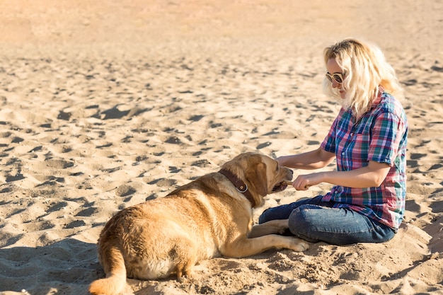 Retrato de uma jovem mulher bonita em óculos de sol, sentado na praia de areia com uma garota de cachorro retriever dourado.