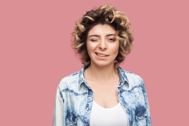 Retrato de uma jovem mulher bonita engraçada com maquiagem e penteado encaracolado em casual camisa azul em pé, piscando e olhando para a câmera com um sorriso. tiro de estúdio interno, isolado no fundo rosa.