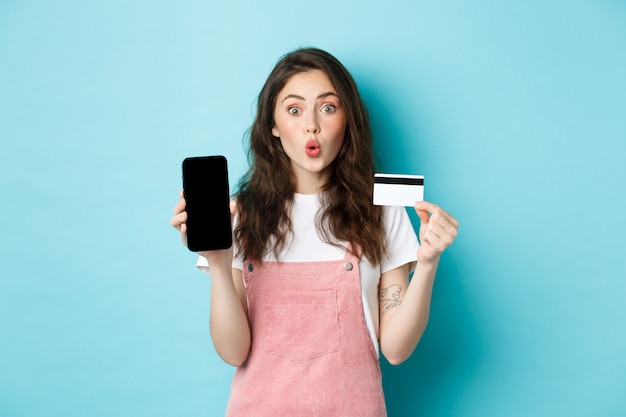 Retrato de uma jovem mulher bonita mostrando cartão de crédito de plástico e tela vazia do smartphone, parece divertido e interessado, mostrando um app interessante, em pé sobre um fundo azul.