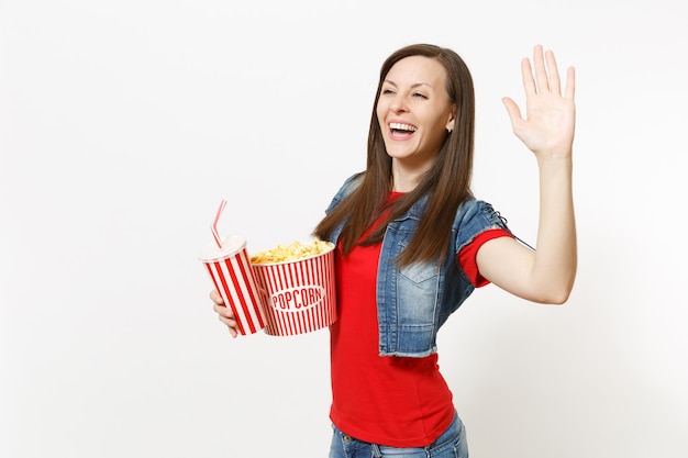 Retrato de uma jovem mulher bonita rindo em roupas casuais, assistindo a um filme, segurando um balde de pipoca, um copo plástico de refrigerante ou cola aparecendo com a palma da mão isolada no fundo branco. Emoções no cinema.