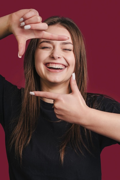 Retrato de uma jovem mulher bonita sorrindo alegremente fazendo um quadro de câmera com os dedos isolados sobre o fundo cor de cereja